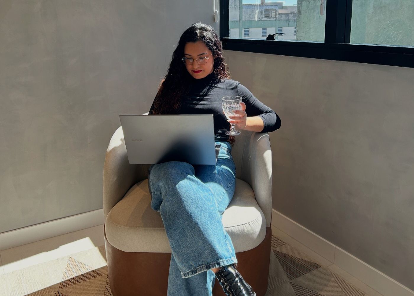 Luana Carolina sentada em uma poltrona ao lado da janela, com o notebook no colo, durante um atendimento online.