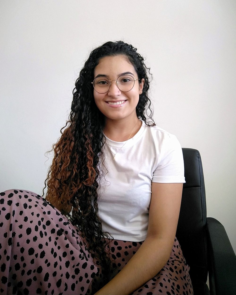 Luana Carolina sorrindo, sentada na poltrona do consultório, de óculos e camiseta branca.